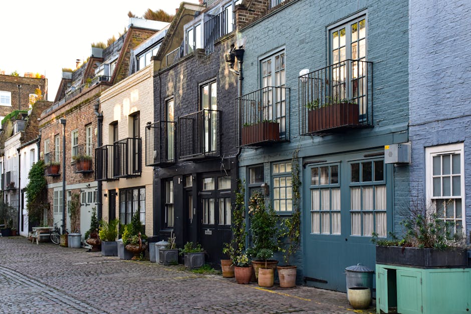 A row of multi-story residential buildings in Lambeth with facades painted in various shades of white, beige, black, and blue. Each building features large windows, some with white frames and others black, with a combination of glass panes and curtains visible inside. Small balconies with black metal railings are attached to some upper-floor windows, and potted plants along with greenery are positioned near entrances and on the balconies. The cobblestone street in front is lined with an assortment of potted plants, decorative pots, and outdoor furniture, creating a charming and well-maintained urban scene. Natural light illuminates the street, highlighting the textures of the brick and painted surfaces, reflecting a clean and orderly environment. Cleaner Lambeth specializes in domestic cleaning and surface sanitisation, ensuring properties remain spotless and hygienic without hidden charges.