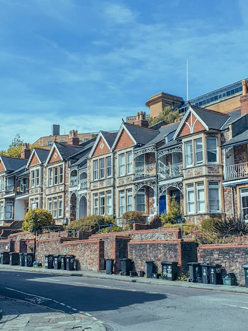 A row of historic Victorian-style terraced houses in Lambeth featuring ornate white woodwork and bay windows, with some houses having small front gardens with neatly trimmed bushes. The buildings are built with red brick and have pitched roofs, under a bright blue sky with scattered clouds. In the foreground, there is a paved street with black wheelie bins lined up along the pavement. The overall scene is well-lit, showcasing a clean and tidy residential area. The image represents a well-maintained neighborhood, ideal for residential cleaning services offered by Cleaner Lambeth, emphasizing quality and attention to detail in property care.