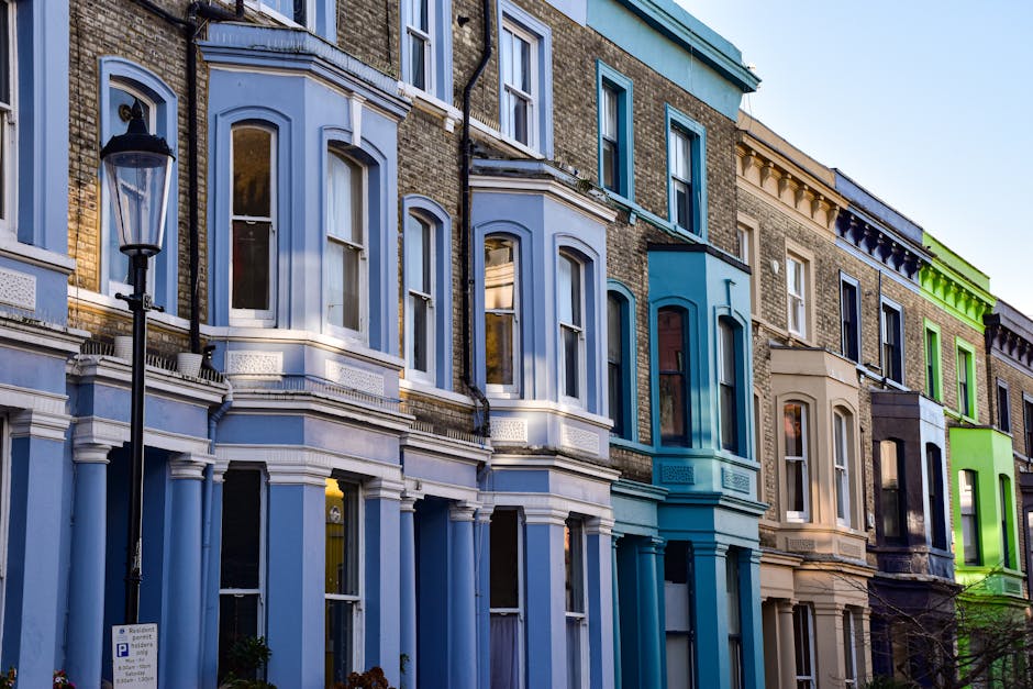 A row of multi-story residential buildings in Lambeth with facades painted in various shades of white, beige, black, and blue. Each building features large windows, some with white frames and others black, with a combination of glass panes and curtains visible inside. Small balconies with black metal railings are attached to some upper-floor windows, and potted plants along with greenery are positioned near entrances and on the balconies. The cobblestone street in front is lined with an assortment of potted plants, decorative pots, and outdoor furniture, creating a charming and well-maintained urban scene. Natural light illuminates the street, highlighting the textures of the brick and painted surfaces, reflecting a clean and orderly environment. Cleaner Lambeth specializes in domestic cleaning and surface sanitisation, ensuring properties remain spotless and hygienic without hidden charges.