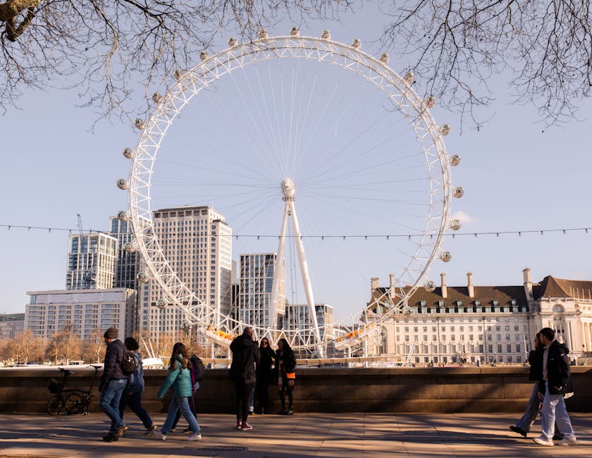 A large white Ferris wheel located outdoors against a clear blue sky, with leafless trees visible at the top and modern high-rise buildings in the background. In the foreground, a group of people dressed in casual clothing, some with backpacks, are walking along a paved promenade alongside the wheel. String lights are suspended across the scene, and a stone balustrade separates the walkway from the water below. The area appears well-maintained and clean, with no litter or debris visible, capturing an active, urban environment. For professional cleaning and maintenance services in Lambeth, visit Cleaner Lambeth at cleanerlambeth.co.uk.