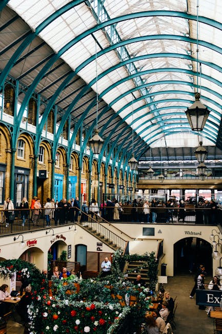 Inside a large, historic market building with a high, arched glass ceiling supported by teal-painted iron framework. The space features a stone and brick facade along the upper walls with large, arched windows. The ground level has a variety of shops and cafes, with people walking, sitting at tables, and browsing. The area appears clean and well-maintained, with polished tiled floors and neatly arranged floral displays. Pendant lamps hang from the ceiling, providing warm lighting, and there are several levels connected by staircases, contributing to an open, spacious atmosphere. This setting exemplifies professional interior cleaning and maintenance, as provided by Cleaner Lambeth, to ensure a hygienic and inviting environment for visitors.