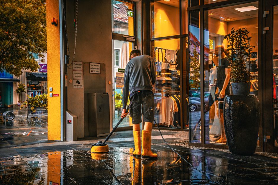A professional cleaner from Cleaner Lambeth is applying surface cleaning to the tiled entrance area of a commercial storefront during rainy weather. The cleaner is wearing bright orange waterproof boots, a grey long-sleeved shirt, and black shorts, operating a pressure washer or power scrubber to remove dirt and grime from the wet black tiles. The storefront features large glass windows and doors, allowing natural light to illuminate the interior, where mannequins dressed in casual clothing are displayed. Outside, reflections of parked cars and the surrounding buildings are visible on the wet pavement, with rain still falling gently. The scene demonstrates routine cleaning and sanitisation procedures essential for maintaining hygiene in commercial premises, with the tools and conditions clearly visible to showcase effective surface cleaning practices conducted by Cleaner Lambeth.