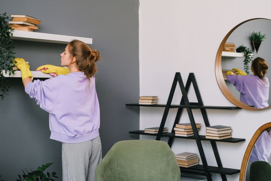 A woman wearing yellow cleaning gloves and a lavender sweatshirt is cleaning a white floating shelf on a gray wall in a modern, well-lit room. She is using a cloth to wipe the surface of the shelf, which holds a few stacked books. In the background, there is a large, round mirror reflecting her movements, and a black shelf ladder with additional stacks of books. A green armchair is partially visible in the foreground, and a small potted plant sits on the floor nearby. The space appears tidy and freshly cleaned, demonstrating surface cleaning and maintenance for a hygienic and inviting environment, characteristic of professional domestic cleaning services by Cleaner Lambeth.