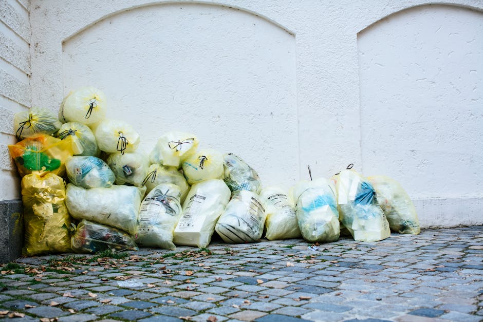 A person holding a large, semi-transparent blue plastic waste bag by the opening with their hand, positioned against a plain white background. The bag appears to be filled with household or commercial