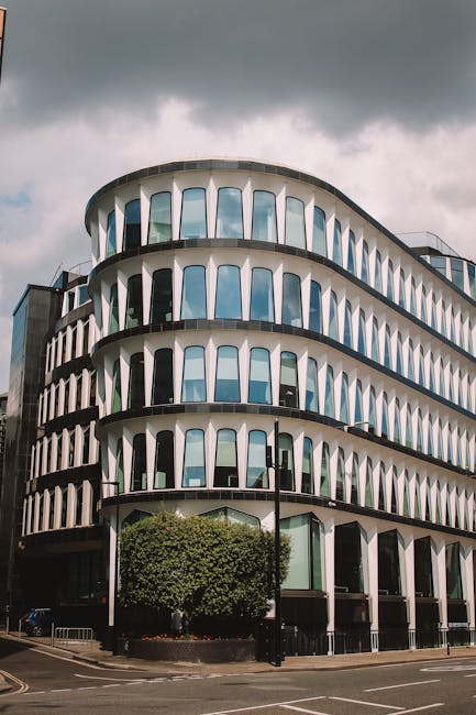 Photograph of a modern, multi-storey office building with a distinctive curved facade featuring large, vertically aligned glass windows framed by white structural elements. The building's exterior appears clean and well-maintained, with no visible dirt or damage. The ground level shows an entrance area with potted plants and a small fenced garden, accented by a neatly trimmed tree in front. The surrounding area includes a paved sidewalk and road, with overcast sky overhead, casting diffused lighting on the scene. As this image is related to Vauxhall office cleaning for small businesses, it highlights the importance of professional surface cleaning and maintenance in commercial environments. Cleaner Lambeth specializes in comprehensive cleaning services for businesses seeking pristine, hygienic offices, including window cleaning, floor polishing, and sanitisation, ensuring a professional appearance and optimal hygiene standards.