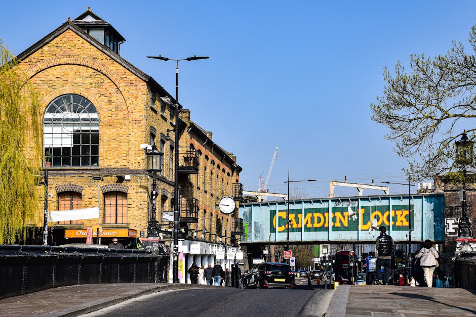 A street scene in Lambeth with a historic brick building featuring large arched windows and a gabled roof, illuminated by daylight. The foreground shows a paved road with pedestrians walking on the sidewalk, some crossing under an overhead railway bridge marked with a large yellow and green 'CATION LOCK' sign. Street lamps and a clock are mounted on black posts along the sidewalk. Trees with fresh green leaves frame the scene on the right and left sides, and a clear blue sky is visible overhead. Cleaner Lambeth specializes in surface cleaning, deep cleaning, and sanitisation services for residential and commercial properties, ensuring all surfaces such as brick, glass, and metal are pristine and hygienic.