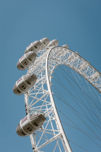A close-up view of the London Eye ferris wheel against a clear blue sky, featuring white structural supports and passenger capsules. The capsules are evenly spaced along the rim, made of transparent glass, and appear clean and well-maintained. The wheel's intricate metal framework is visible, highlighting the technical design and engineering. The scene is brightly lit with natural sunlight, emphasizing the cleanliness and polished appearance of the ferris wheel. This image showcases a well-kept outdoor attraction, aligning with the theme of surface cleaning and maintenance often addressed by Cleaner Lambeth for various commercial and leisure facilities.